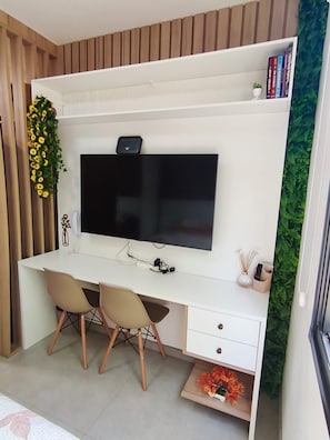 Habitació moderna amb un escriptori blanc, dues cadires, un televisor de pantalla plana i una petita estanteria amb llibres i objectes decoratius.