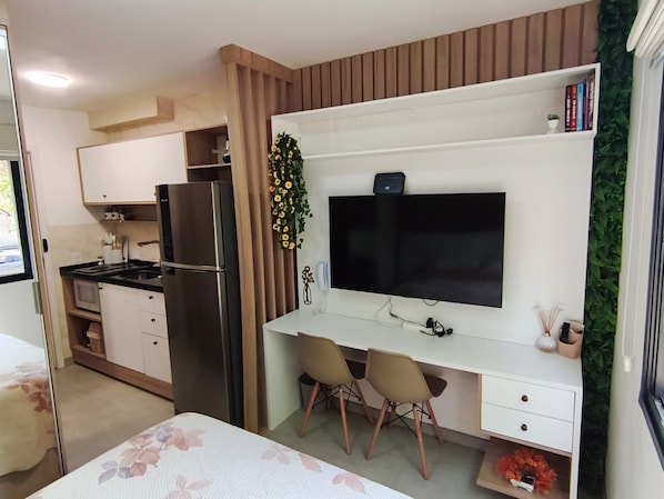 Un espai habitable compacte amb una cuineta, un televisor de pantalla plana penjat a la paret, un petit escriptori amb dues cadires i un llit amb teles florals.