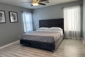 Un dormitorio con cama, ventilador de techo y dos ventanas con cortinas.