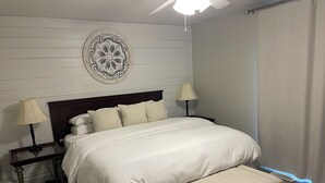 A bedroom with a bed, two bedside tables, a nightstand, a decorative wall plate, a ceiling fan, and a window with curtains.