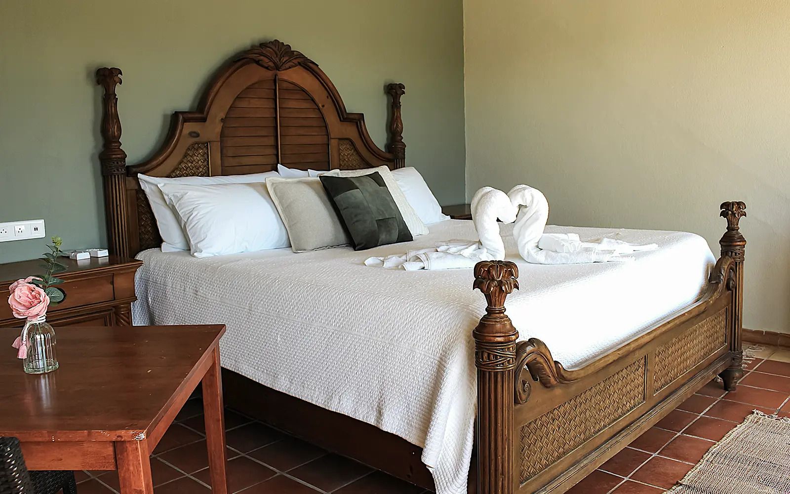 Una cama bien hecha con cabecera de madera, una mesita de noche con un florero y una alfombra en el suelo.