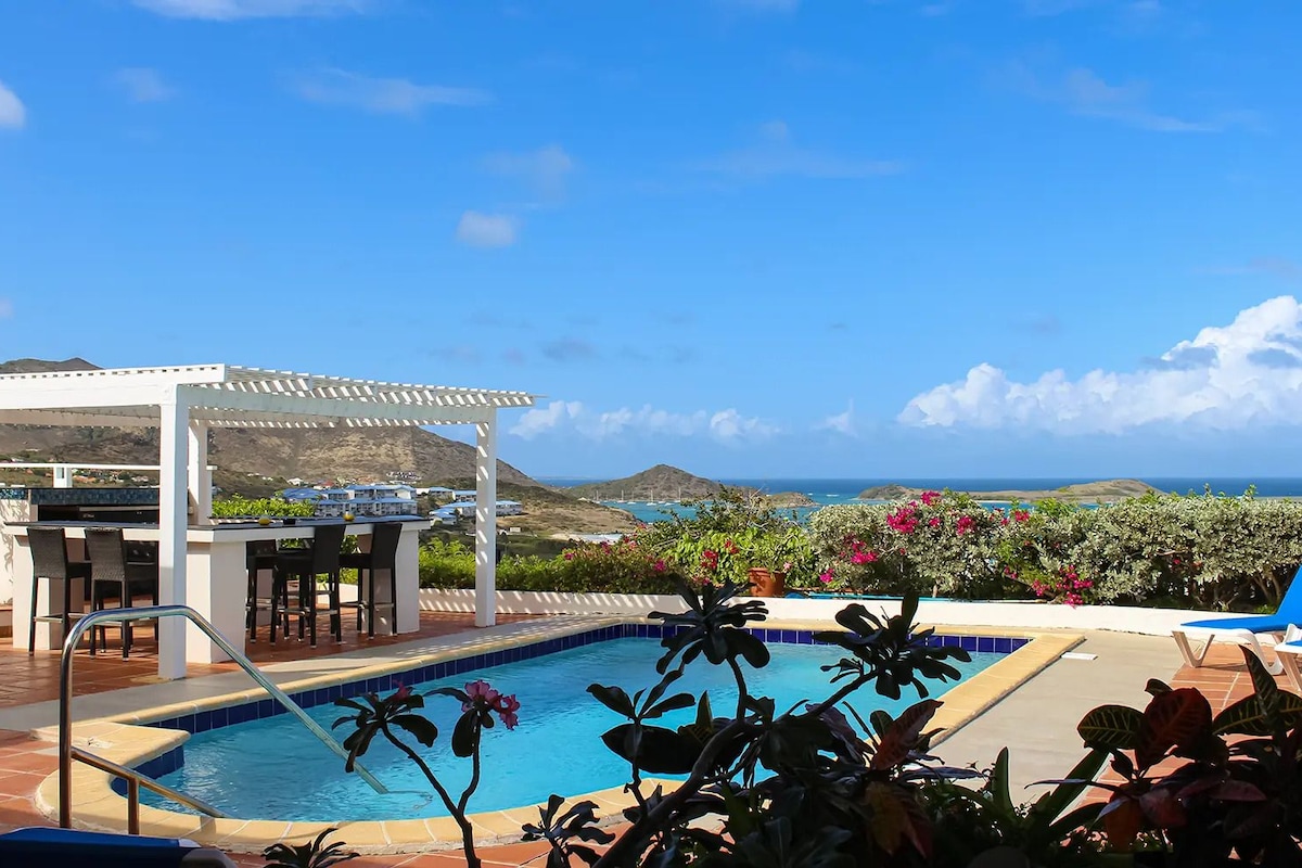 A pool area with a pergola, outdoor seating, and a view of the ocean.