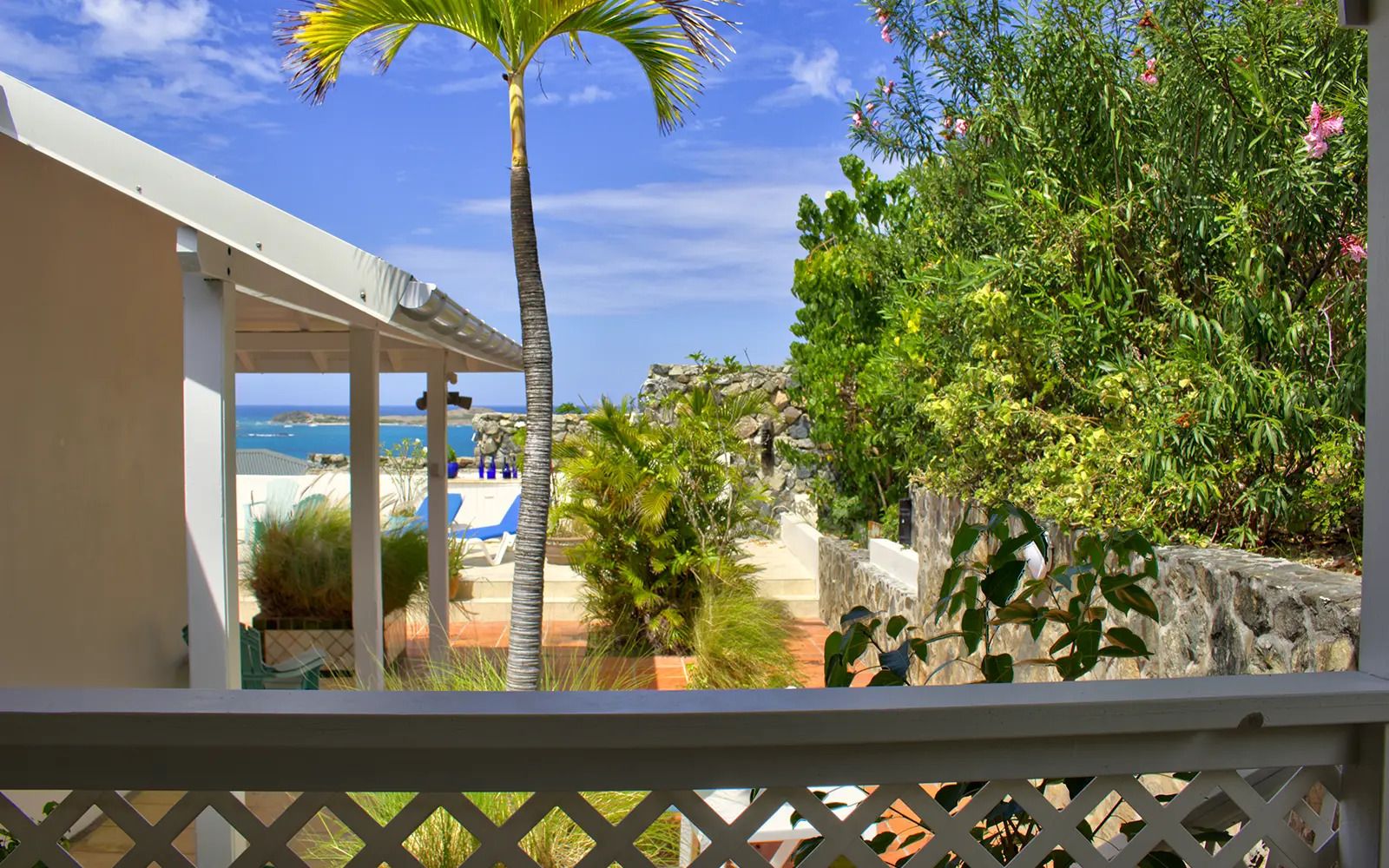 Vista desde un balcón sobre un área de piscina con una palmera, un muro de piedra y vistas al mar.