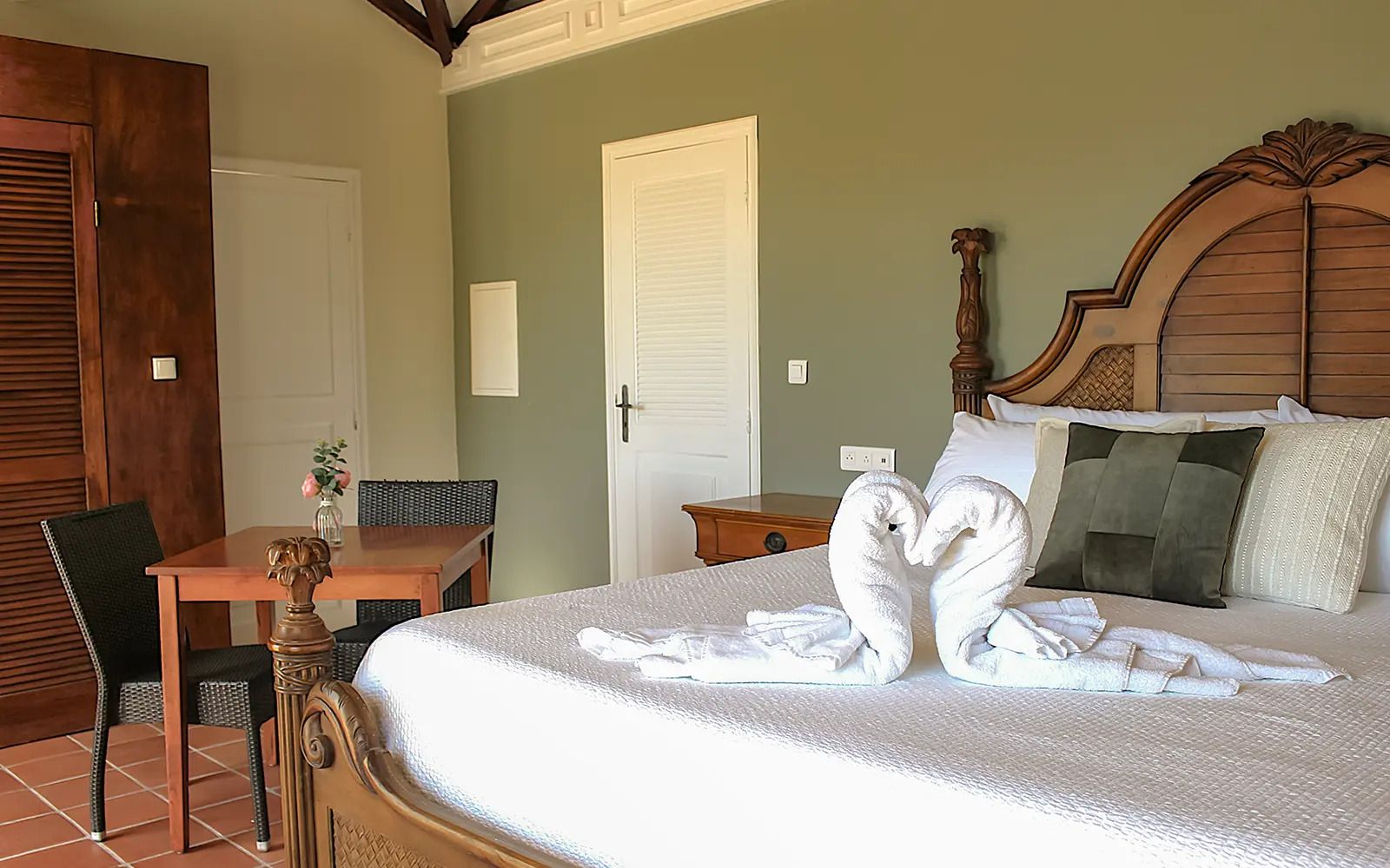 Habitación de hotel con una cama, un cabecero de madera, una mesa de comedor con sillas y una mesita con un florero.
