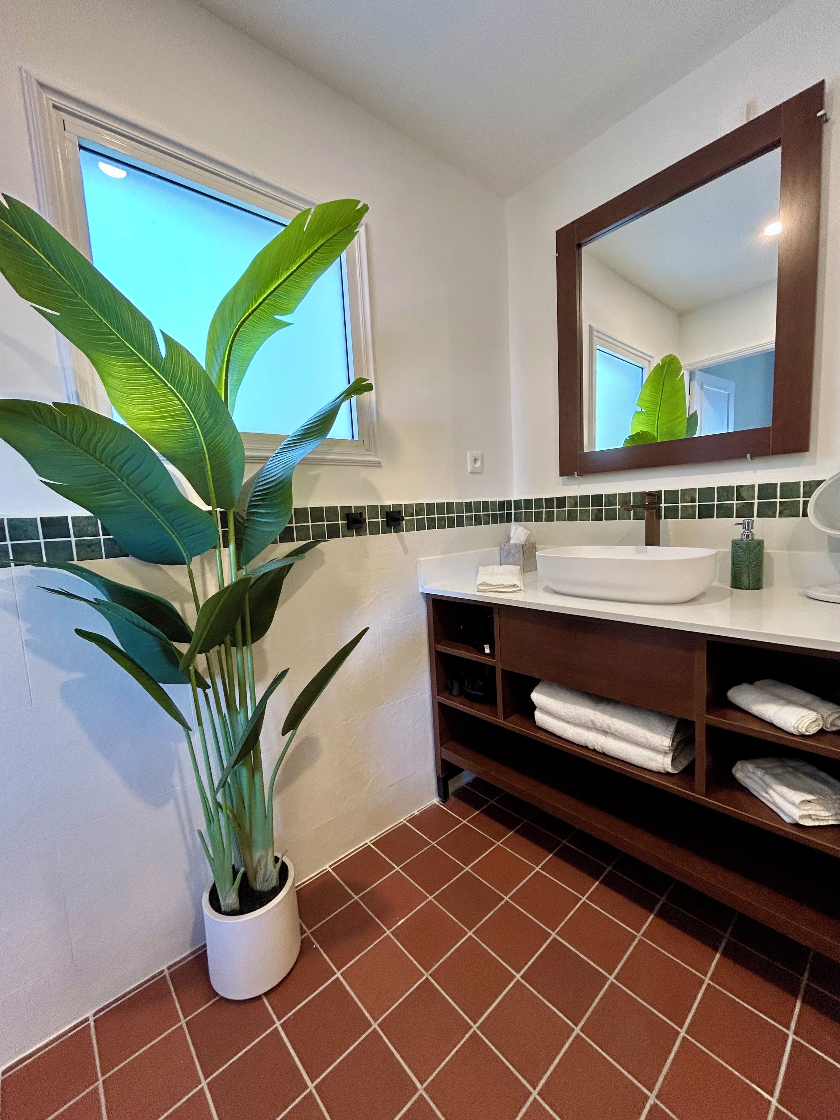 Un baño con lavamanos blanco, espejo, planta en maceta y suelo de baldosas.