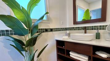 Un baño con lavamanos blanco, espejo, planta en maceta y suelo de baldosas.