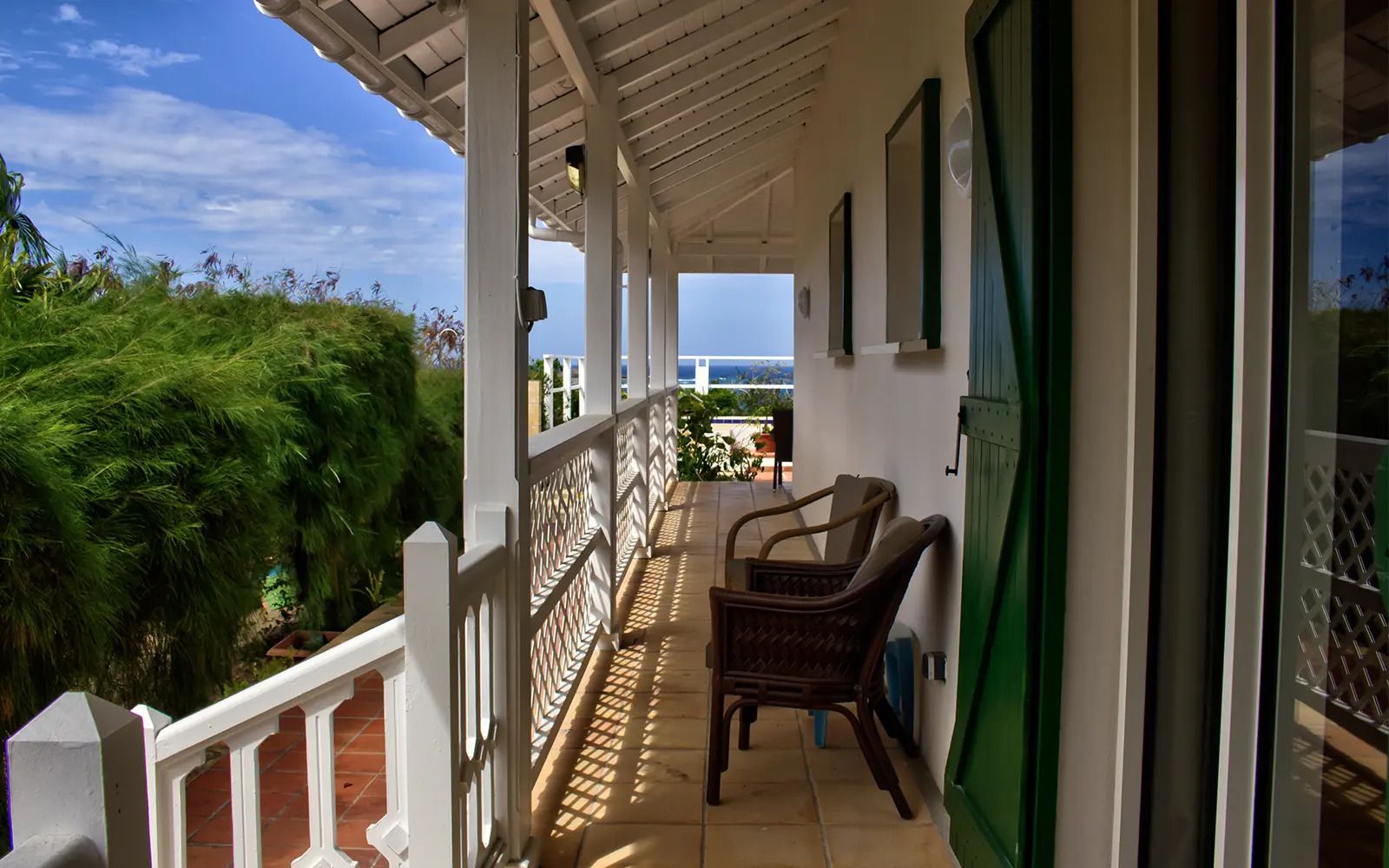 Un balcon abrité avec vue sur l’océan, une chaise en osier et une porte verte.
