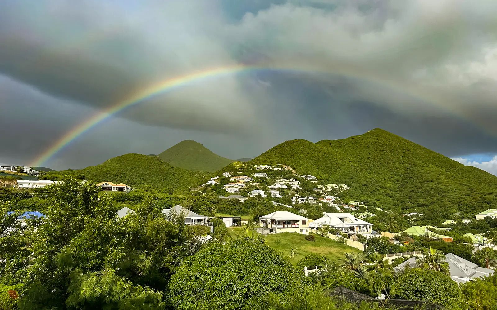 A residential area with houses on a hillside, a mountain in the background, and a rainbow.