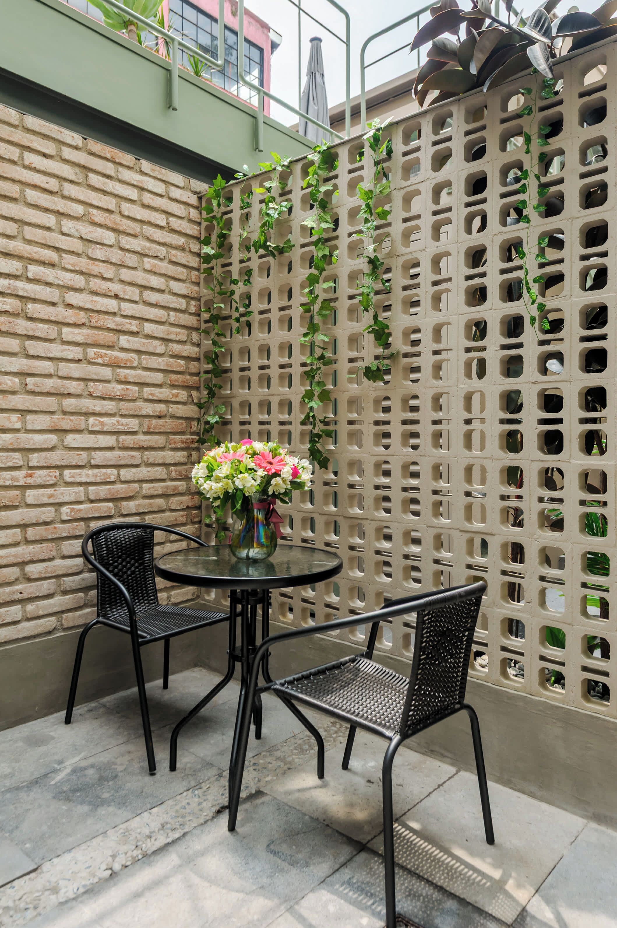 A patio area with a brick wall, a green trellis with climbing plants, and two black metal chairs around a glass table with a vase of flowers.
