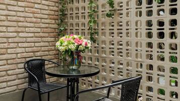 A patio area with a brick wall, a green trellis with climbing plants, and two black metal chairs around a glass table with a vase of flowers.