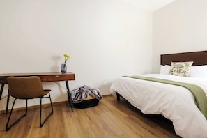 Habitación de hotel con un escritorio de madera, una silla, una cama con ropa blanca y un plaid verde, y un pequeño florero con flores en el escritorio.