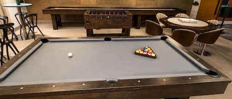 Uma mesa de bilhar com bola branca e um conjunto de bolas coloridas, cercada por cadeiras e uma mesa de futebol de botão.
