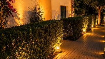 Un chemin pavé mène à un bâtiment doté d’une porte, qui est entouré de verdure et éclairé par des luminaires au sol.