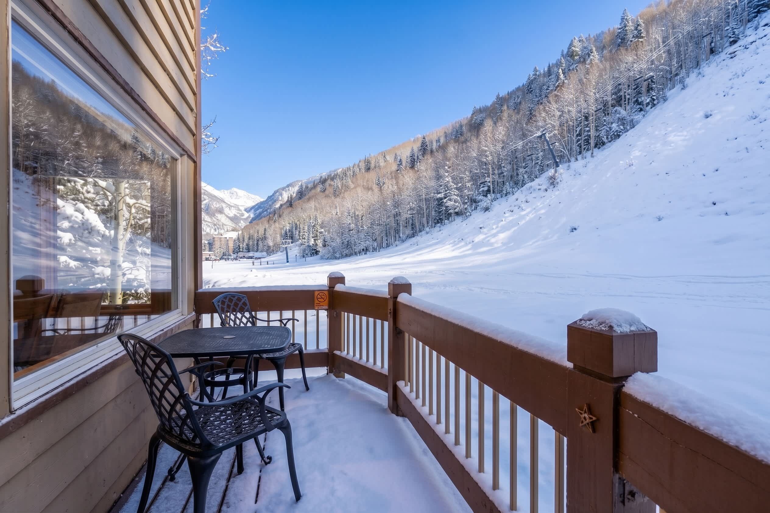 Um deque coberto de neve com uma mesa e cadeiras, com vista para uma paisagem montanhosa nevada.