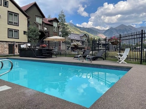 Ein Schwimmbecken mit Liegestühlen und Grillplatz, umzäunt und von Gebäuden gesäumt, mit Blick auf eine Berglandschaft im Hintergrund.
