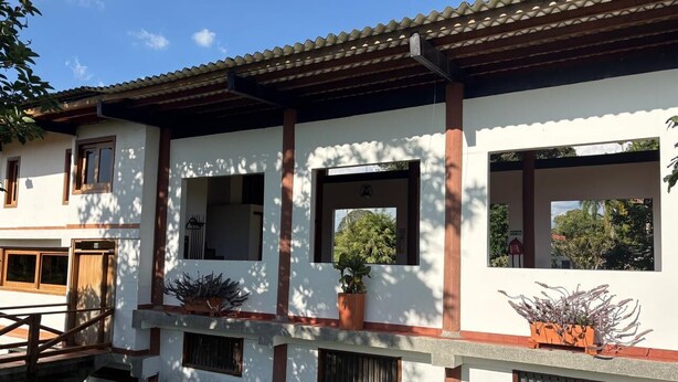 Ein zweistöckiges Gebäude mit weißer Fassade, braunen Verzierungen und einem Ziegeldach. Im zweiten Stock befinden sich Fenster, und ein Balkon mit Blumentöpfen.