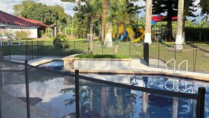 Ein Schwimmbecken mit Glasbarriere, umgeben von einem eingezäunten Bereich mit Palmen und einem Spielplatz im Hintergrund.