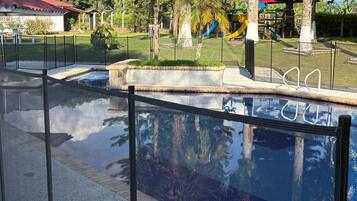 A swimming pool with a glass barrier, surrounded by a fenced area with palm trees and a playground in the background.