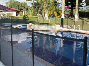 A swimming pool with a glass barrier, surrounded by a fenced area with palm trees and a playground in the background. - Vrbo Property (rural)