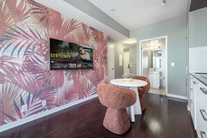 Ein modernes Interieur mit einem wandmontierten Fernseher, einem runden Tisch mit zwei Stühlen und einem durch einen offenen Durchgang einsehbarer Küchenbereich.