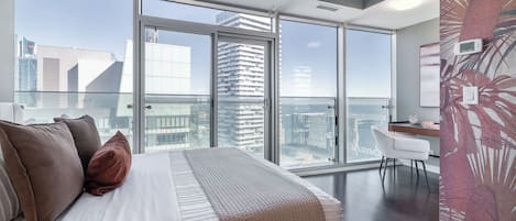 Et moderne soveværelse med en stor seng, et skrivebord og udsigt over byen gennem gulv til loft-vinduer.