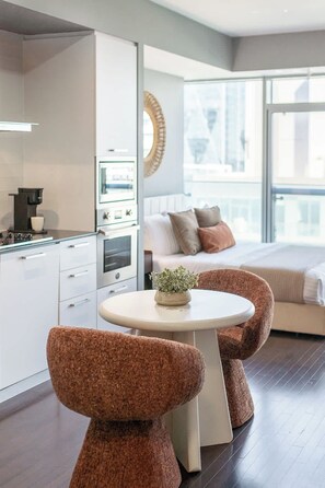 Een moderne hotelkamer met kitchenette, een ronde eettafel, twee gestoffeerde fauteuils, een bed met kussens en een groot raam.
