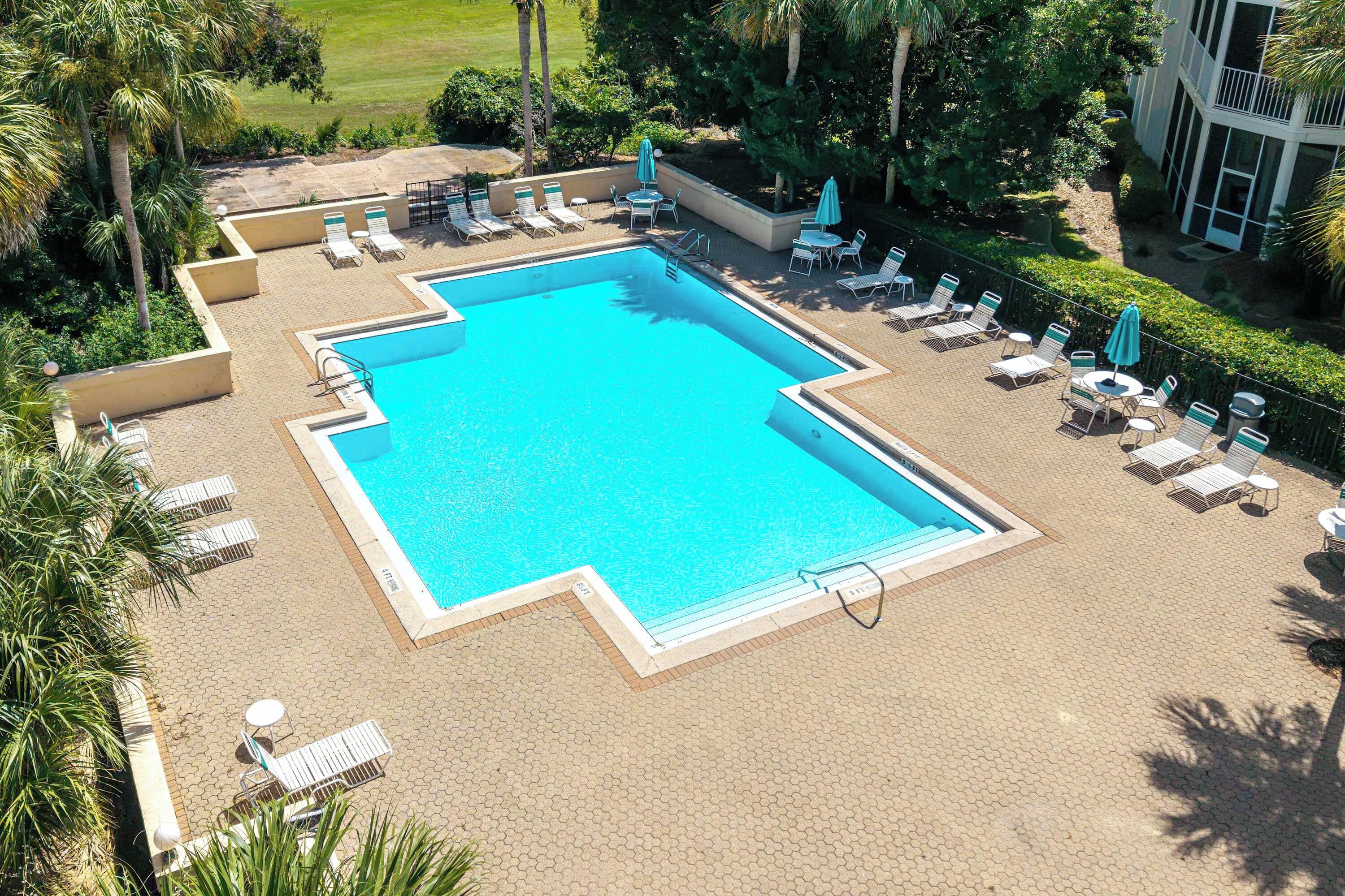 Une piscine rectangulaire entourée de chaises longues et de parasols, avec un jardin aménagé et un bâtiment en arrière-plan.