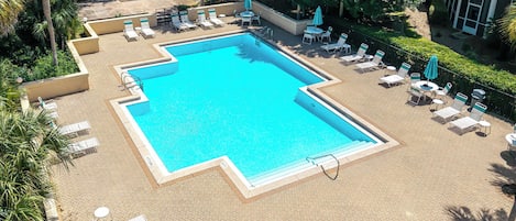 Piscina retangular rodeada por espreguiçadeiras e guarda-sóis, com um jardim ajardinado e um edifício ao fundo.