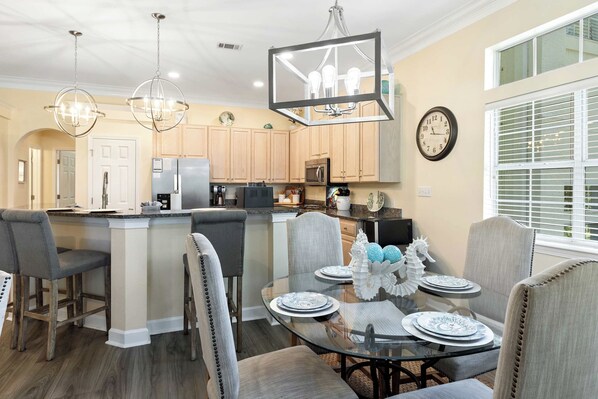 A modern kitchen with wooden cabinets, a central island, and a dining area with a glass table and chairs. - Luxury Golf Cart & View, Steps to Beach,Pool,Tram (Miramar Beach)