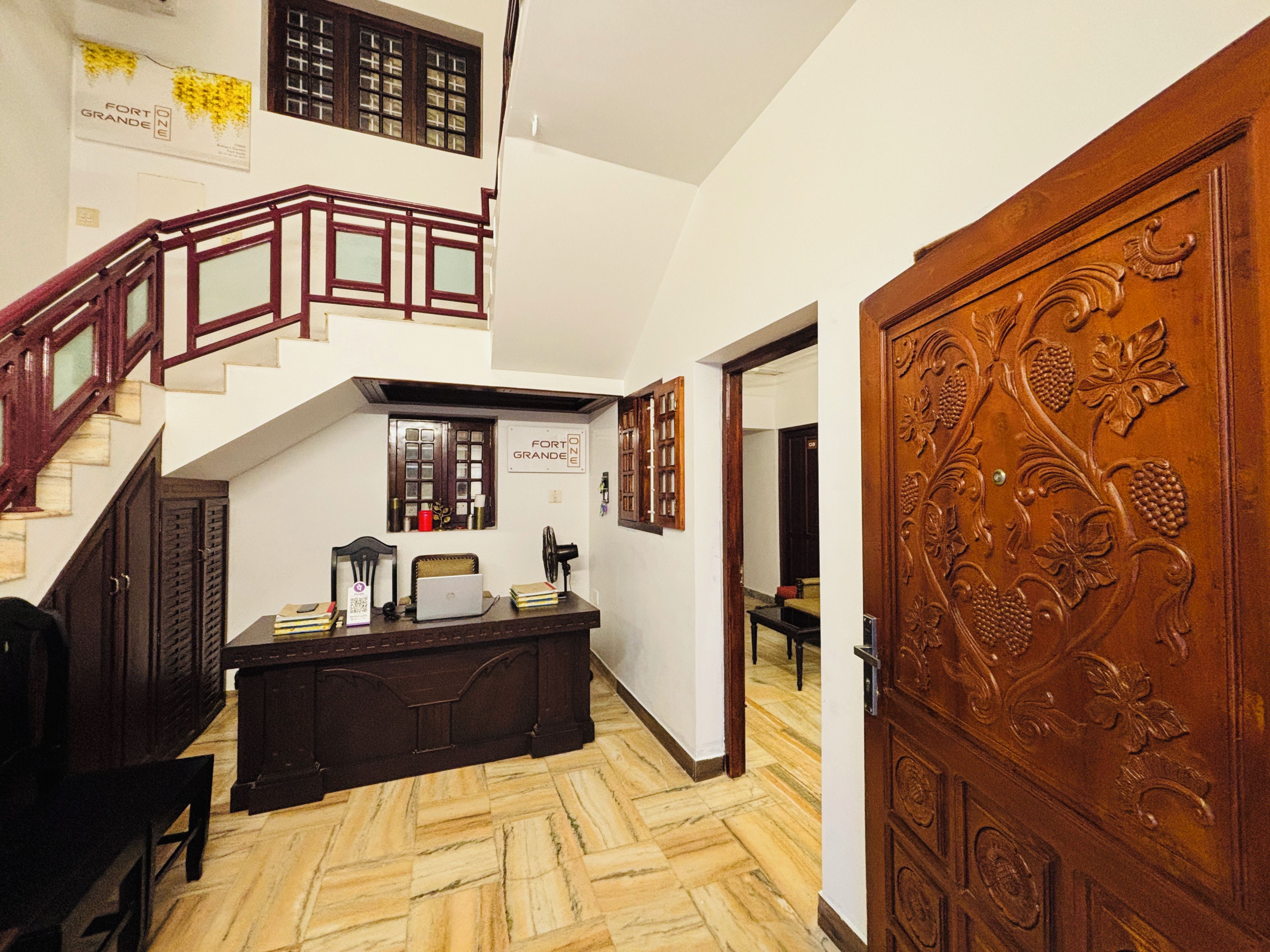 A staircase with wooden railings leading to an upper floor, a desk with a laptop, and a large wooden door with intricate carvings.