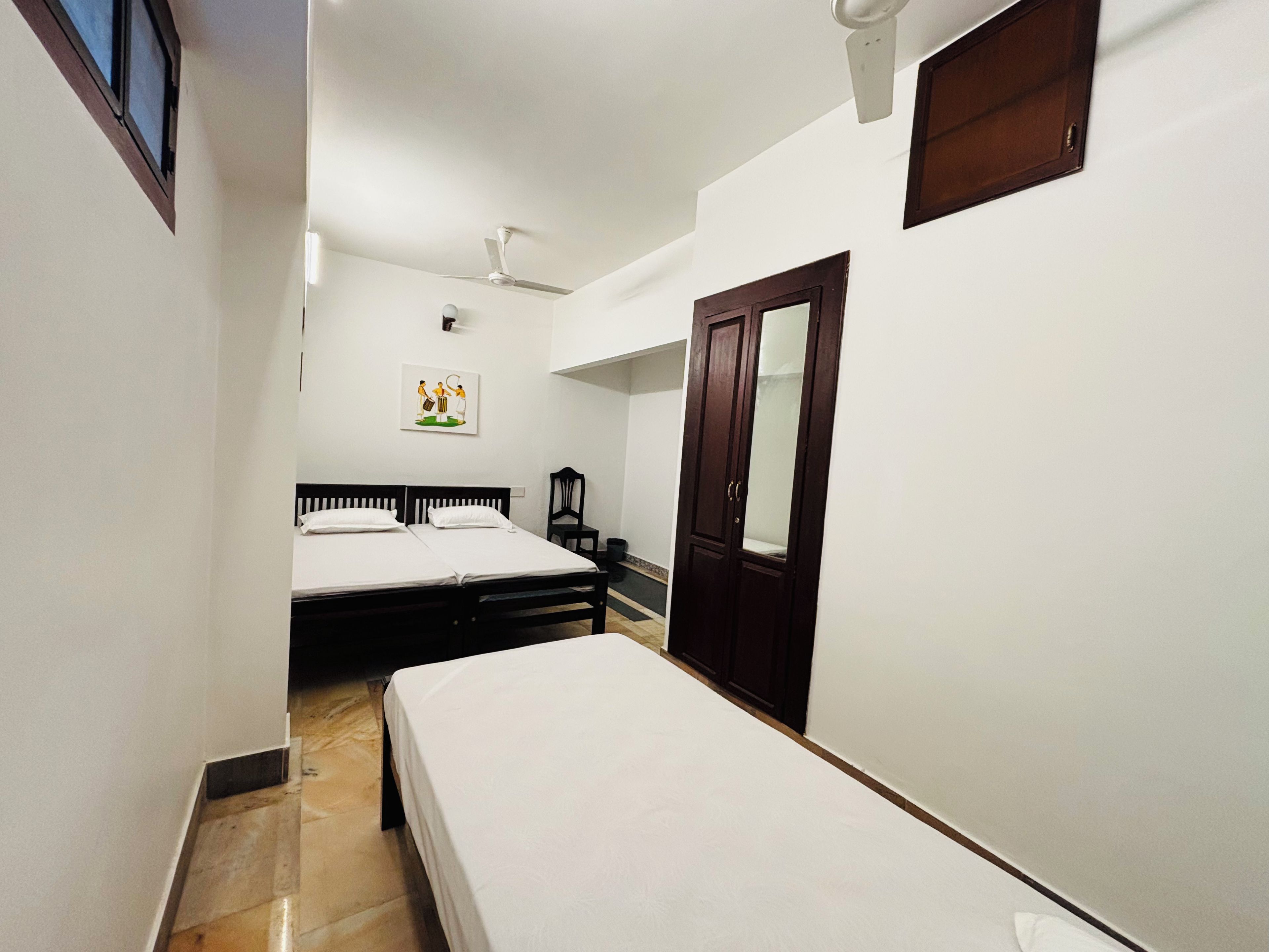 Una habitación de hotel estrecha con dos camas, un ventilador de techo, una puerta y un ventanal pequeño.