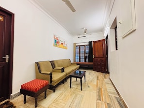 A room with a sofa, a small table, a bench, a ceiling fan, a window with curtains, and a door. - Fort Grande ONE (Kochi)