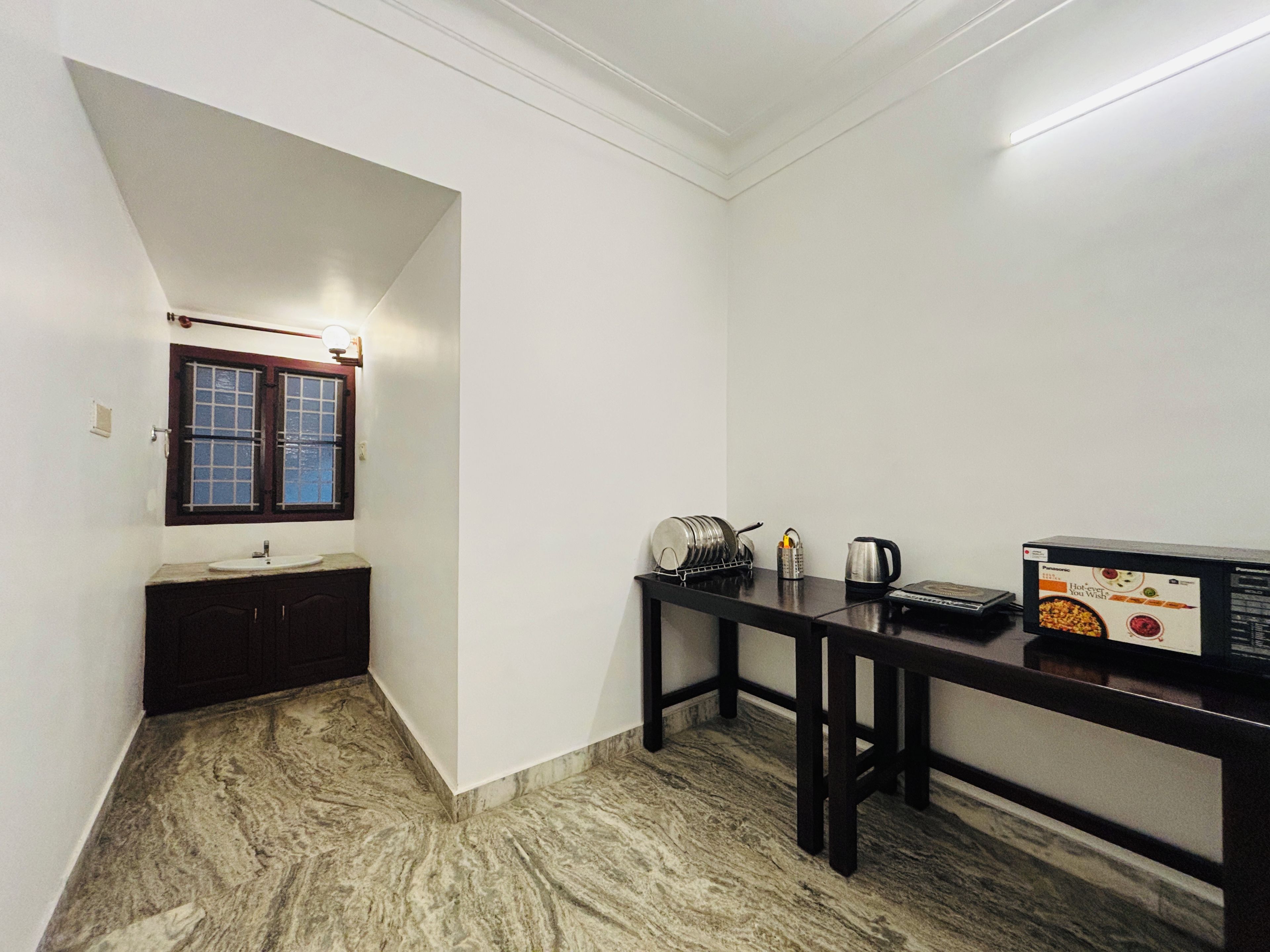 A compact kitchen with a marble floor, a sink, a window with a wooden frame, and a table with a kettle and a box of hot water tea.