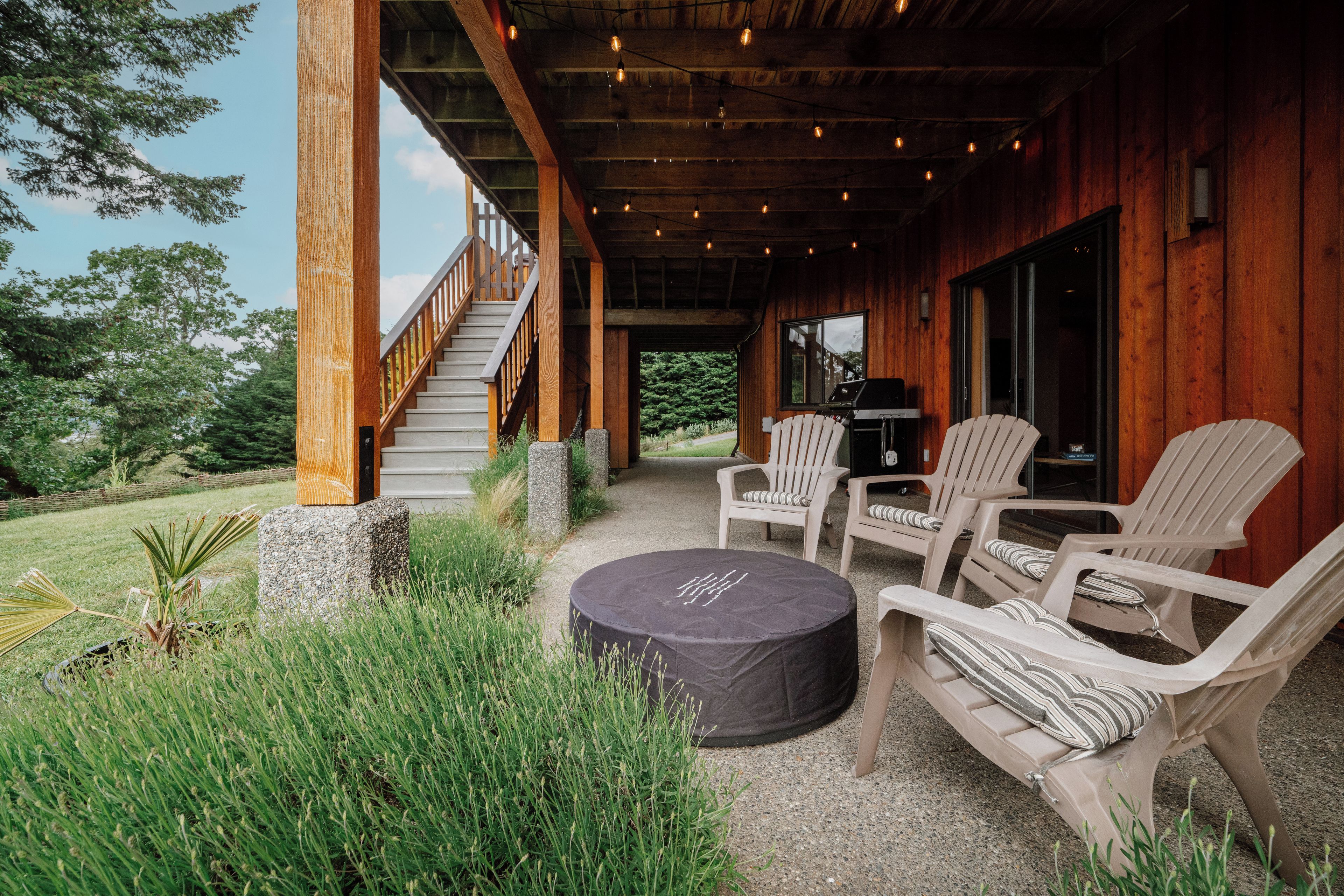 A covered outdoor seating area with Adirondack chairs and a round table, a staircase leading to an upper level, and a view of the surrounding greenery.