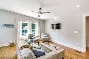 A living room with a sofa set, two armchairs, a flat-screen TV mounted on the wall, and a ceiling fan.