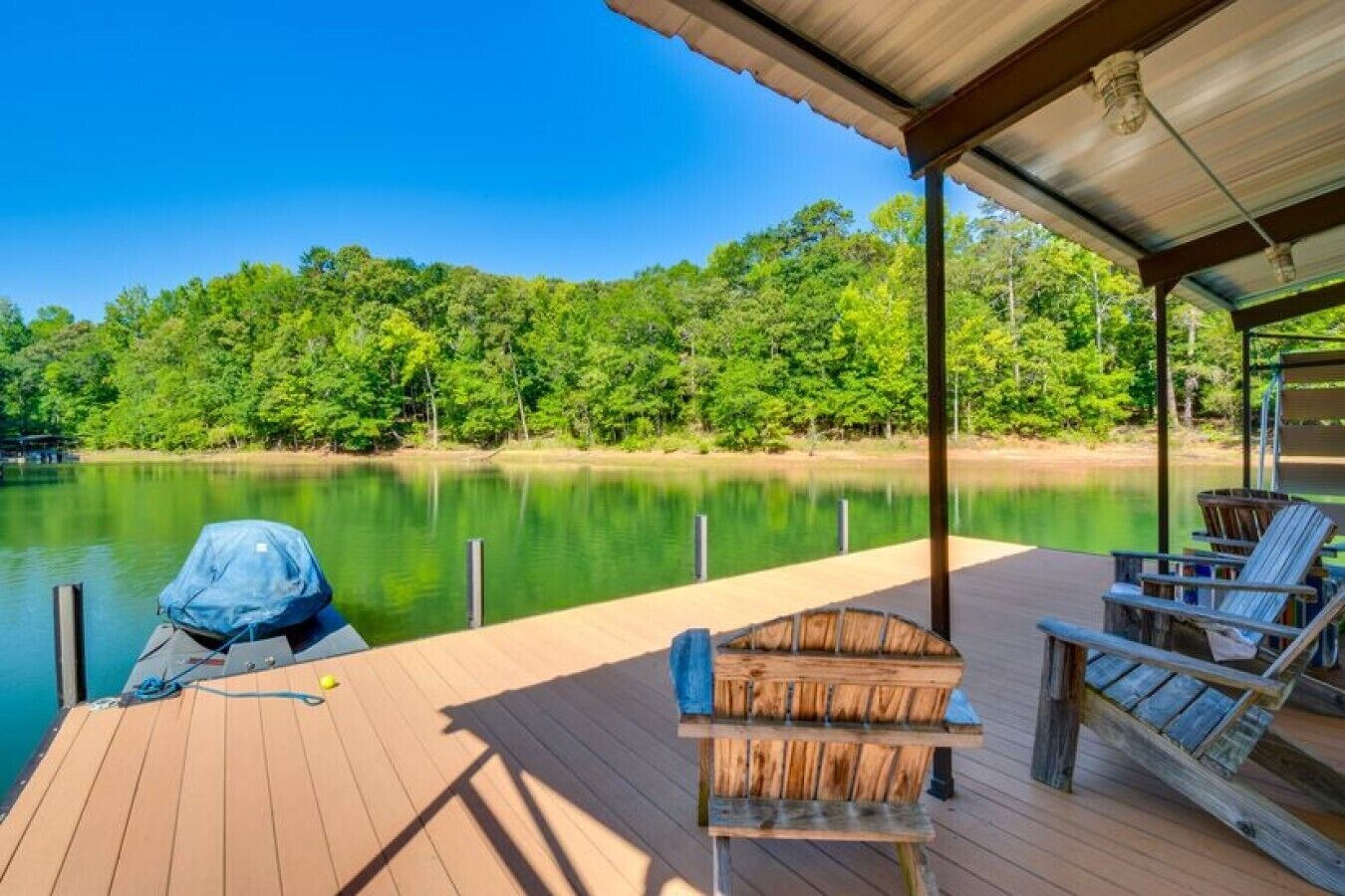 Eine Holzterrasse mit überdachtem Bereich, einem Boot und Blick auf einen See mit Bäumen im Hintergrund.