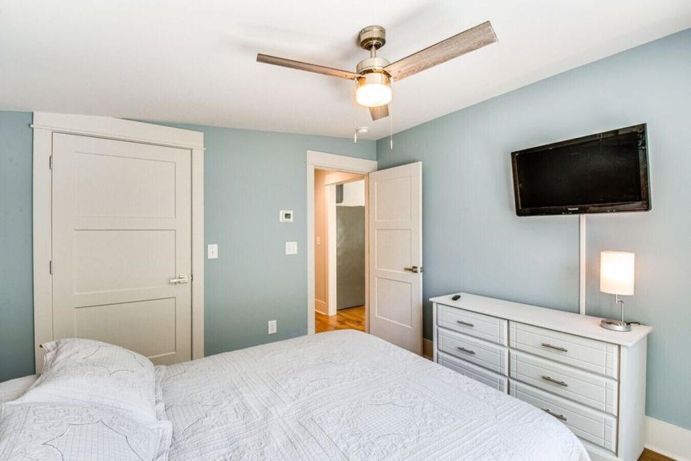 A bedroom with a bed, a dresser, a ceiling fan, and a television on the wall.