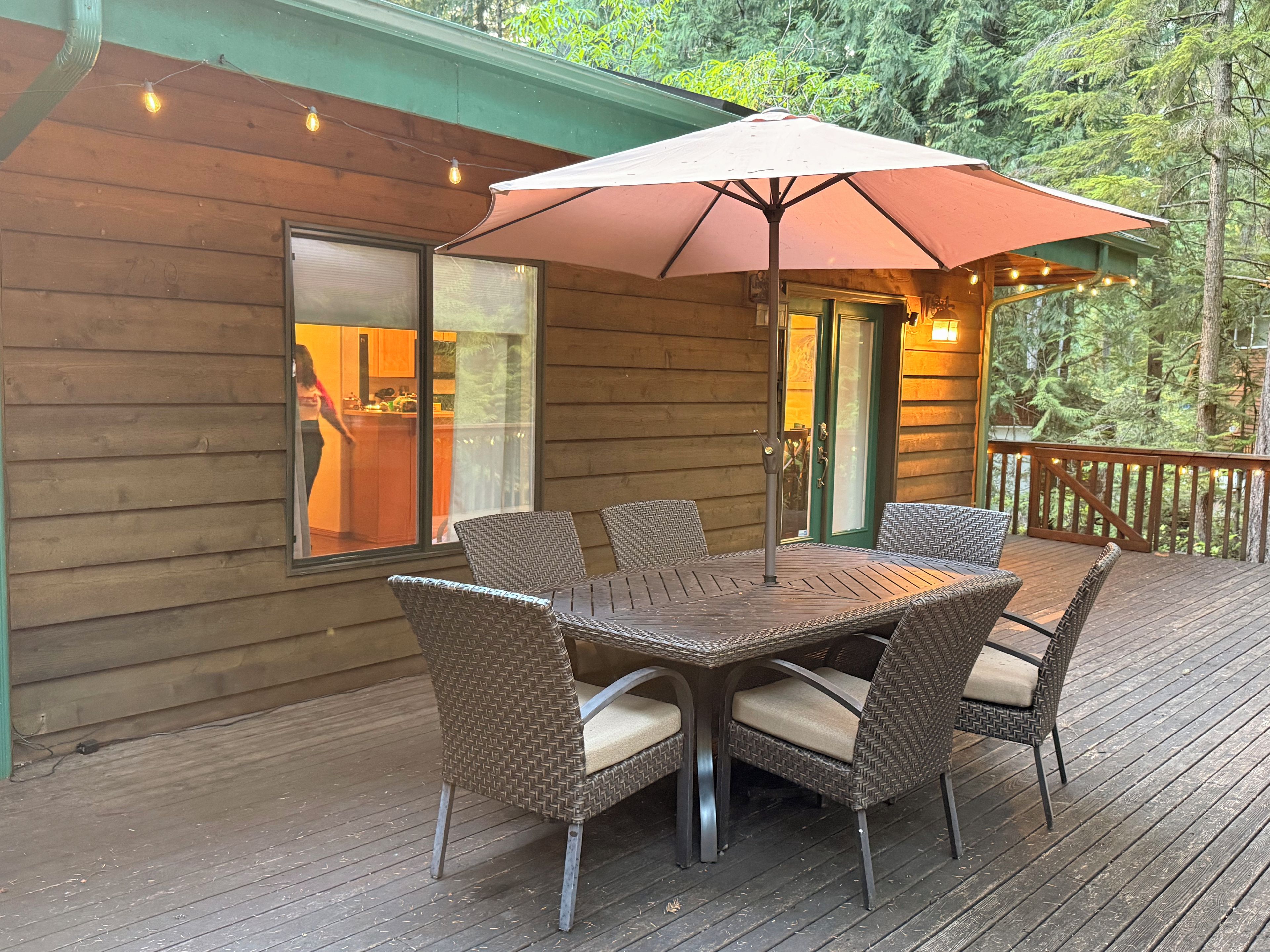 Une terrasse en bois avec une table et des chaises, un parasol et une vue sur une cuisine à travers une fenêtre.