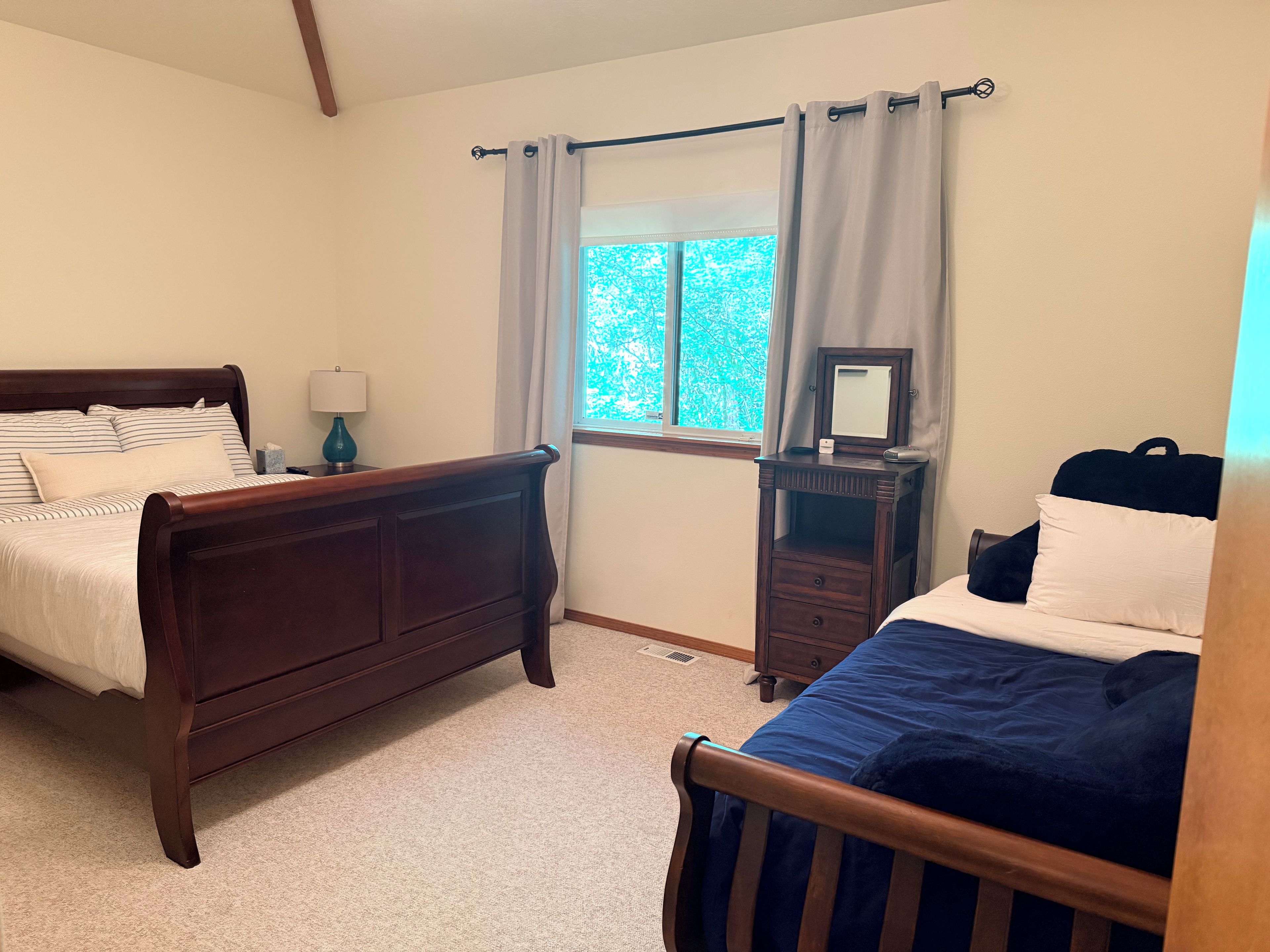 A bedroom with two beds, a nightstand, a dresser, a window with curtains, and a view of greenery outside.