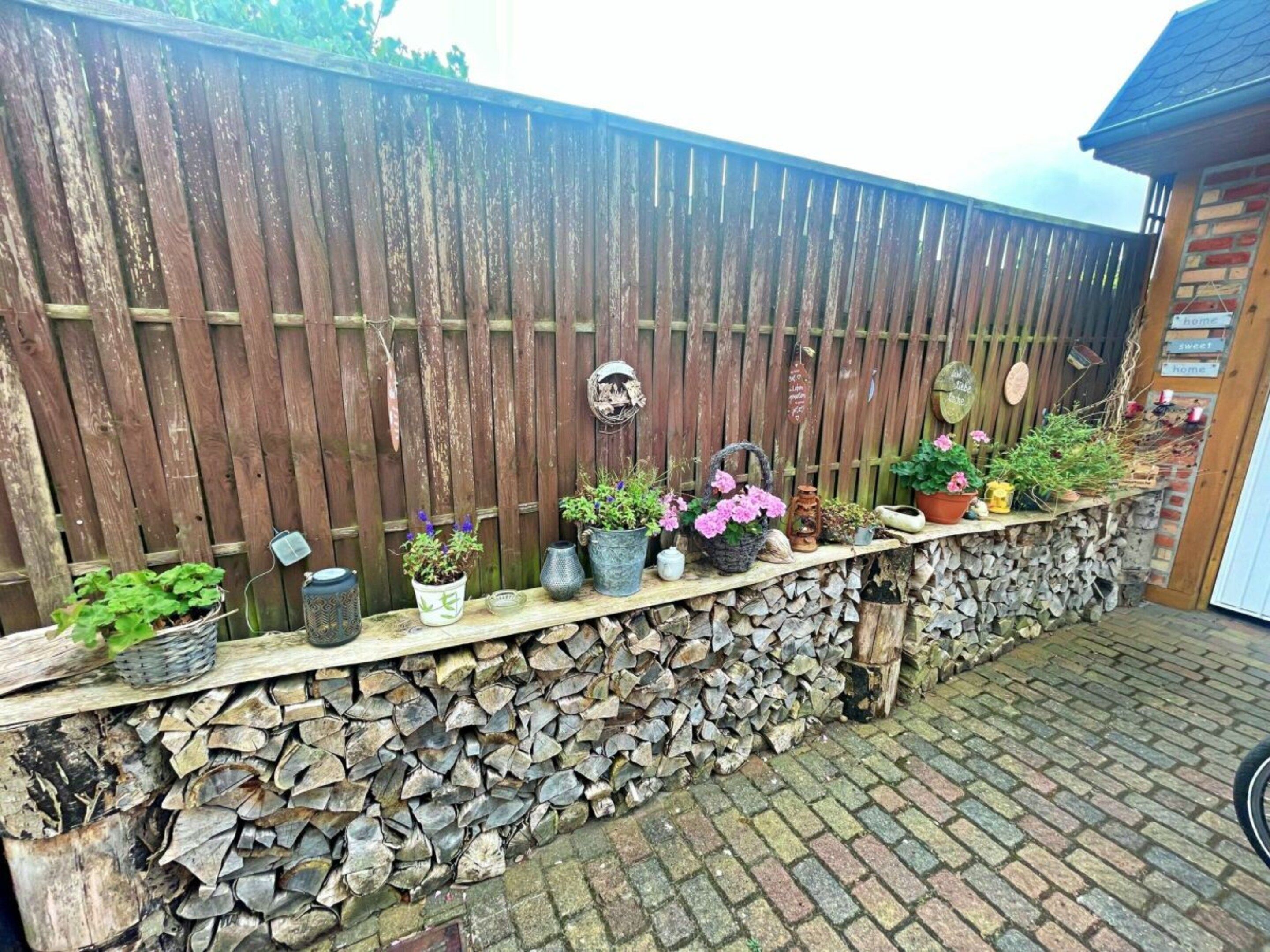 Une clôture en bois, adossée à un mur en pierre, et une étagère en bois où sont disposées des plantes en pot et des objets de décoration de jardin.