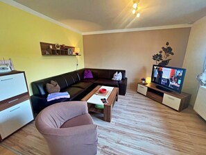 Ein Wohnzimmer mit einer Couch, einem Couchtisch, einem Fernseher und einem Schreibtisch mit einem Computer.