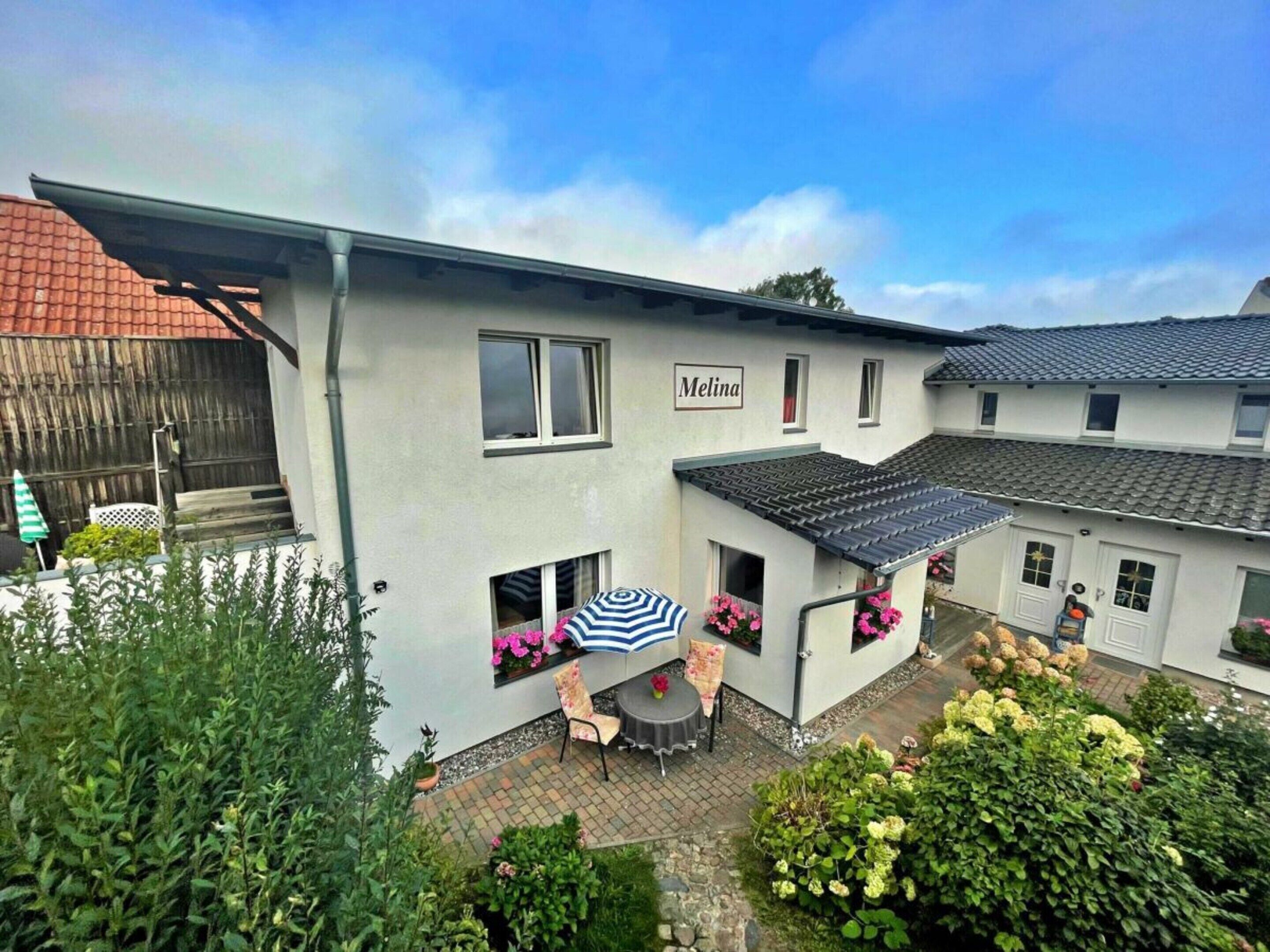 Un bâtiment à deux étages avec une enseigne « Melina » sur le côté. On y trouve une terrasse avec une table et des chaises, ainsi qu’un espace de repos couvert avec un parasol à rayures.