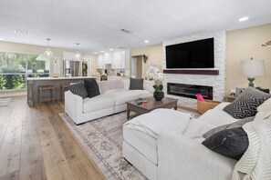 Een moderne woonkamer met een open haard, een keuken op de achtergrond en een grote televisie aan de wand.