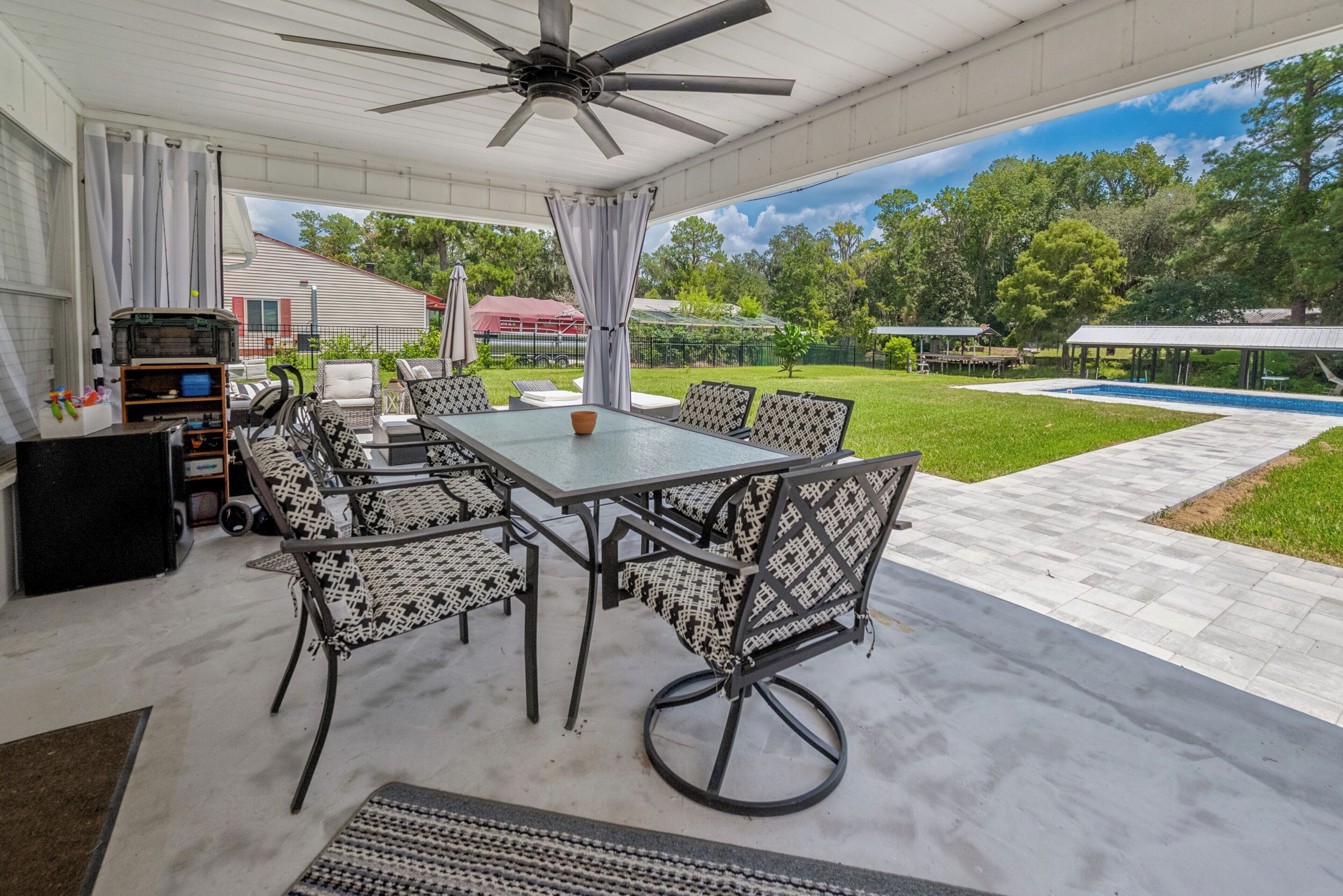 An outdoor patio with a table and chairs, a ceiling fan, and a view of a swimming pool and greenery.