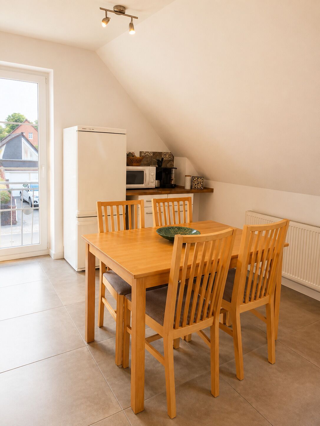 Un'area pranzo con un tavolo e sedie in legno, un frigorifero bianco, un forno a microonde e una finestra con vista su una strada residenziale.