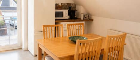 A dining area with a wooden table and chairs, a white refrigerator, a microwave, and a window overlooking a residential street.