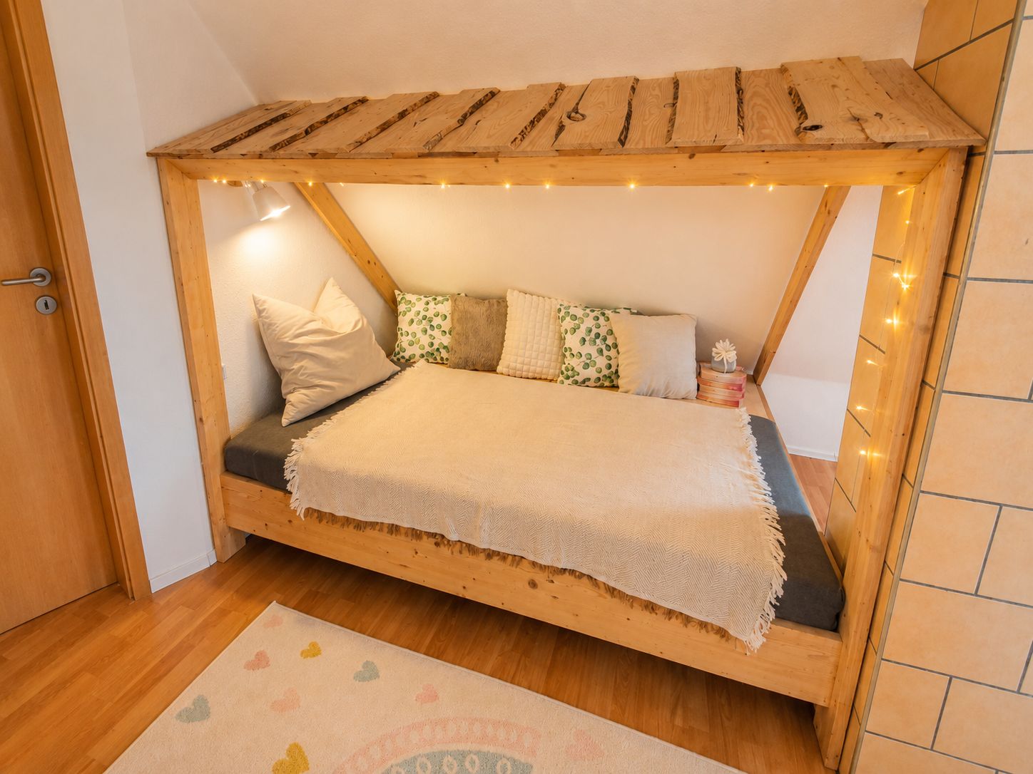 Een houten bed met schuine balken, een bedlampje en een decoratief kussen.