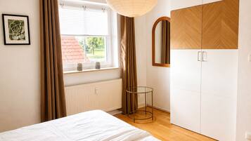 A bedroom with a bed, a window with curtains, a radiator, a bedside table, and a wardrobe.