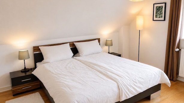 A neatly made bed with white linens, two bedside tables with lamps, a floor lamp, a framed picture on the wall, and a door with curtains. - Meerbeck Vacation Rental (Meerbeck)