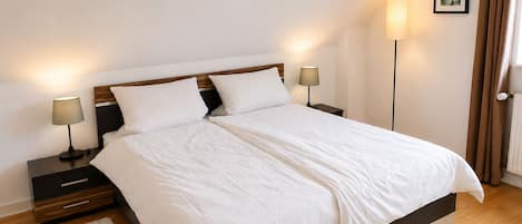 A neatly made bed with white linens, two bedside tables with lamps, a floor lamp, a framed picture on the wall, and a door with curtains.